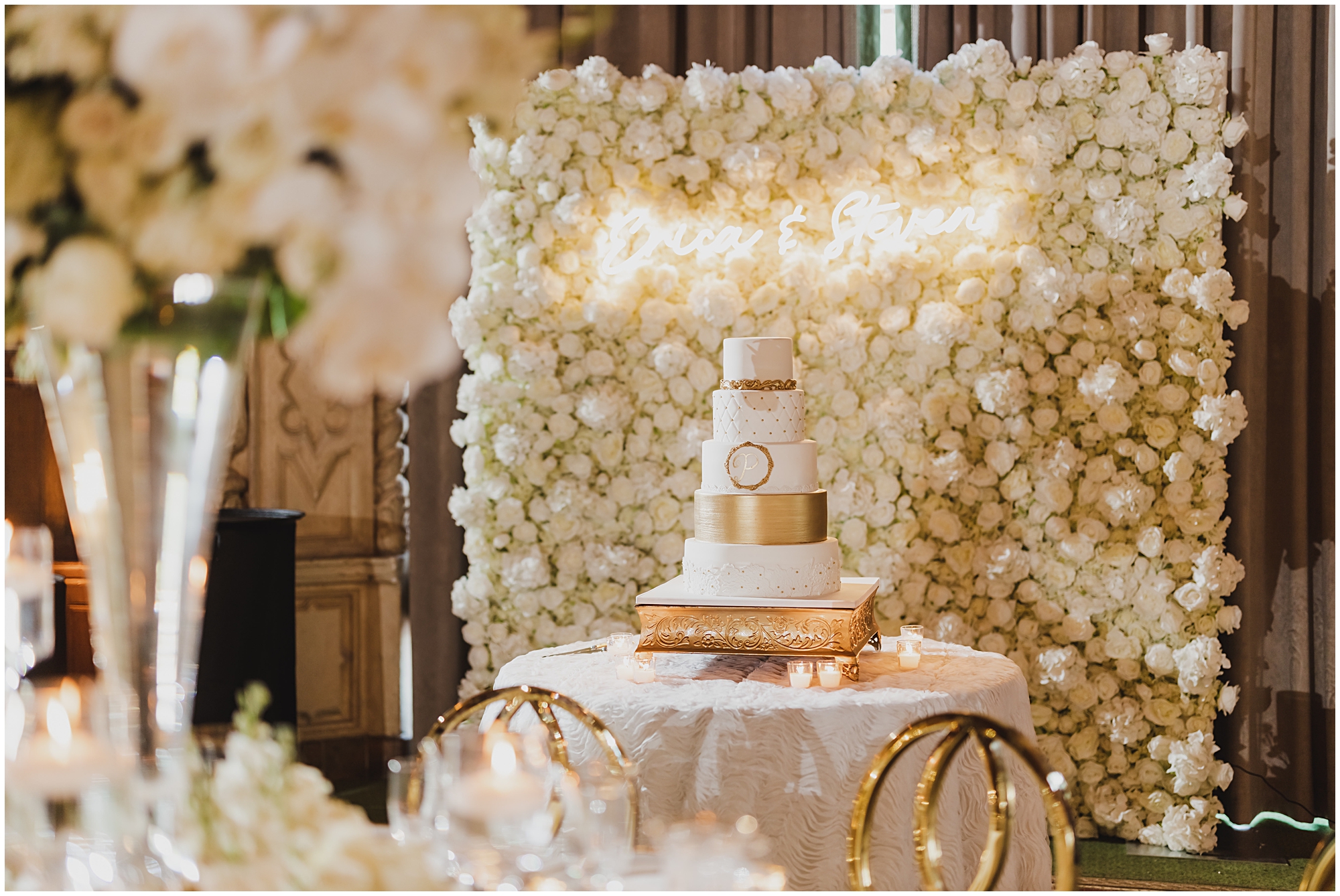 White and gold cake in front of a flower wall under a neon sign.
